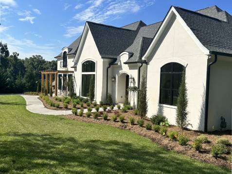 New Project - 2025-02-11T170743.481 A modern kitchen features a central island with a stovetop and range hood, two clear barstools, white cabinets, stainless steel appliances, and wooden flooring. An orchid plant decorates the island.