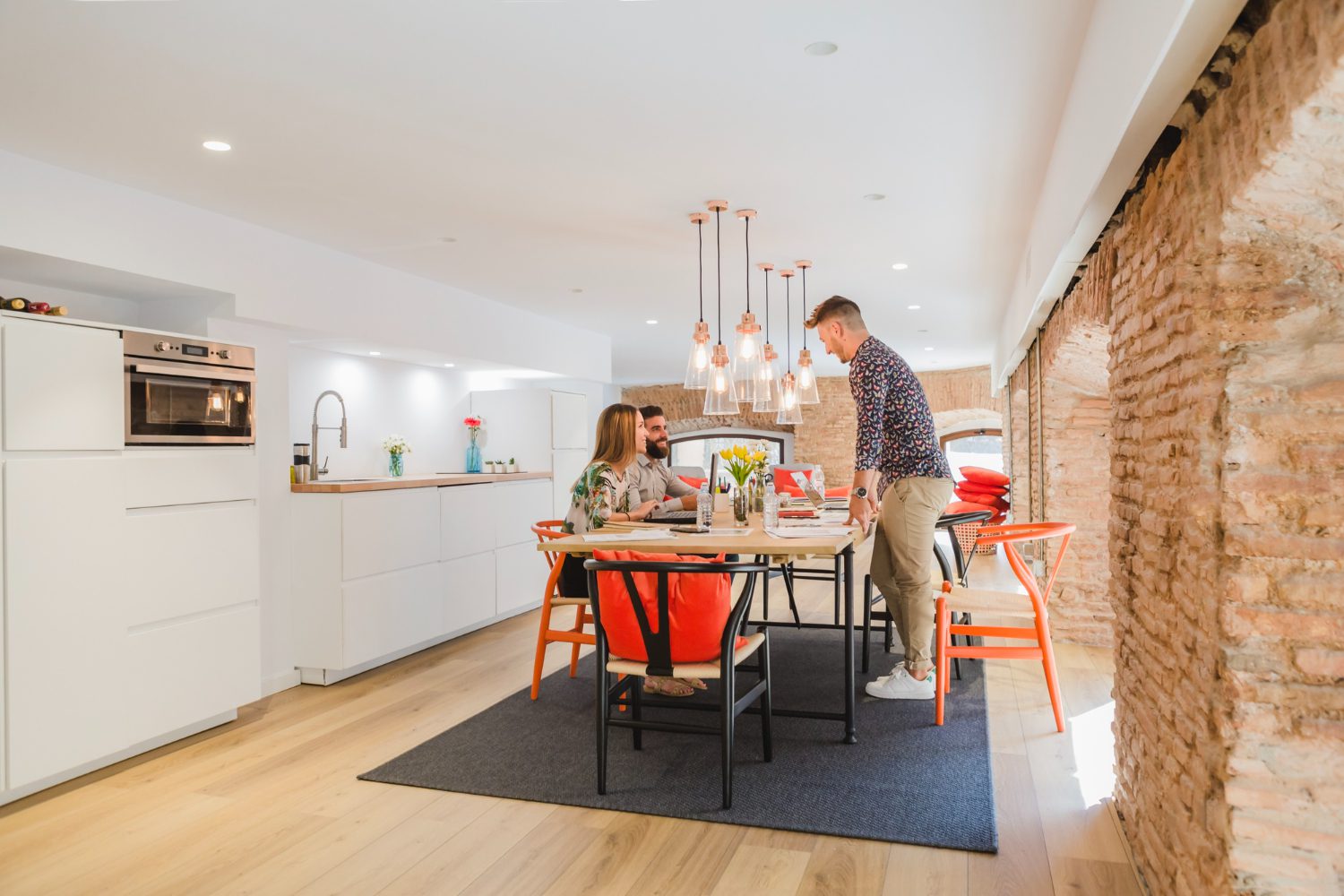 2147668768 Three people are gathered around a dining table in a modern kitchen with exposed brick walls and contemporary lighting.